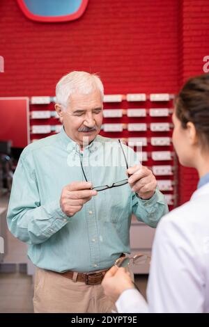 Senior man looking at his eyeglasses while standing in front of young female ophthalmologist, consulting with her and trying on new eyewear Stock Photo