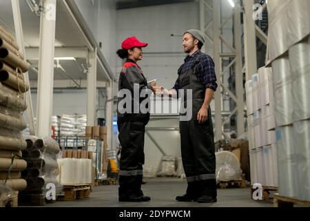 Young male professional in overalls and checkered shirt shaking hand of his female colleague with digital tablet during work in warehouse Stock Photo