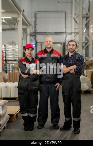 Group of successful male and female professionals of large contemporary plant standing in front of camera in aisle between packed materials Stock Photo