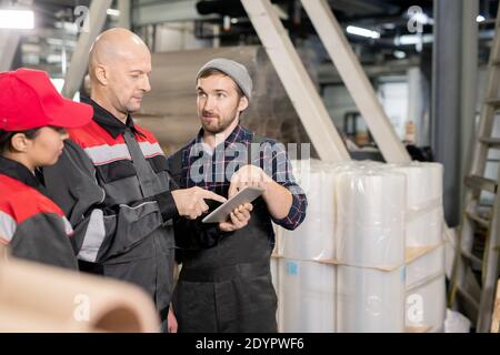 Serious bald mature foreman and one of his subordinates in workwear pointing at tablet display during presentation of new materials Stock Photo