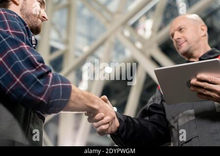Bald mature foreman with digital tablet greeting his young subordinate by handshake before meeting in warehouse or factory environment Stock Photo