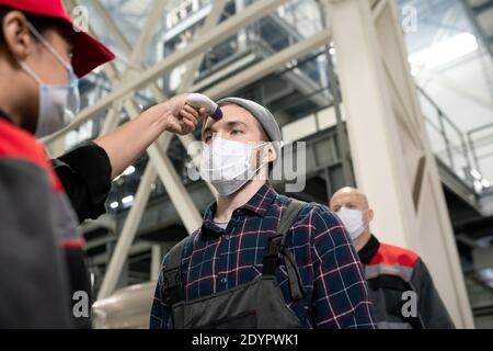 Young female factory worker in protective mask measuring body temperature of one of engineers against huge construction Stock Photo