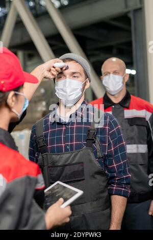 Young medical worker in uniform and protective mask measuring and fixing in database body temperature of factory workers in the morning Stock Photo
