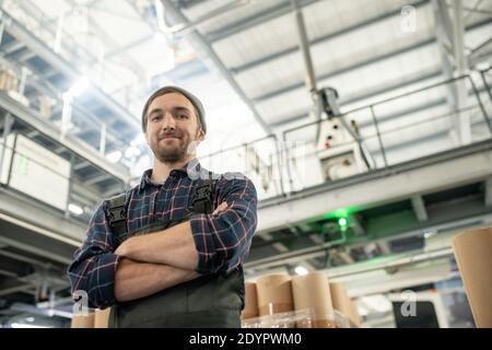 Young successful engineer of large factory in shirt, beanie and coveralls standing with crossed arms in front of camera against huge construction Stock Photo