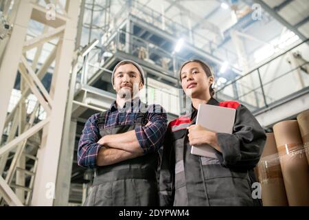 Young successful male and female engineers of large plant standing in front of camera and looking at you against huge technical construction Stock Photo