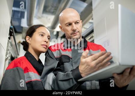 Serious bald mature foreman in workwear holding laptop and pointing at data on display while making presentation to his young female subordinate Stock Photo