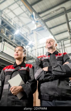Contemporary confident male and female workers of large modern factory standing in front of camera against warehouse environment Stock Photo