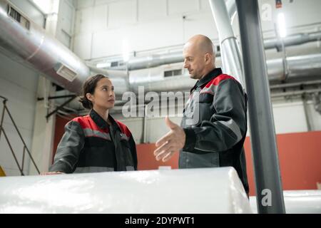Young female factory worker and her male colleague in workwear standing by huge packed roll in warehouse and consulting before unpacking it Stock Photo