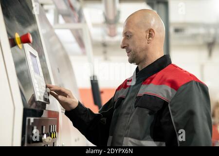 Bald mature worker of contemporary large polymer processing factory pressing button on control panel while resetting industrial machine Stock Photo
