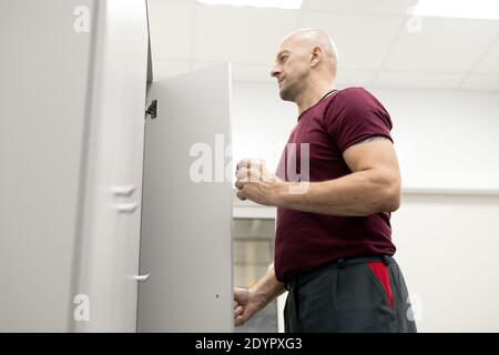 Side view of bald serious mature worker of modern factory having drink while opening door of locker with his workwear and other stuff Stock Photo