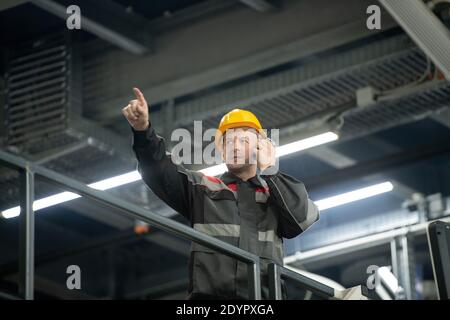 Confident mature foreman with hardhat on head and walkie talkie by his ear pointing forwards during work in large polymer processing factory Stock Photo