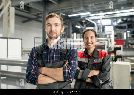 Two young successful workers of polymer processing factory crossing arms by chest while standing in front of camera in large workshop Stock Photo