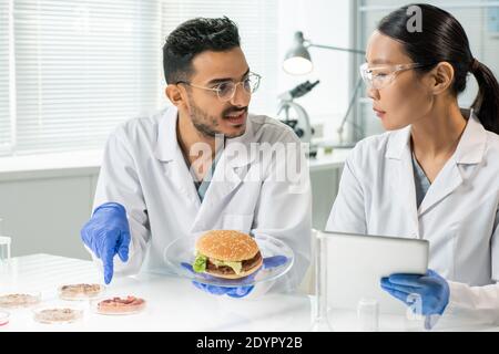 Gloved young man with hamburger on plate pointing at one of samples of raw vegetable meat while describing its characteristics to colleague Stock Photo