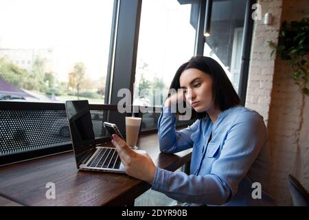 Tired female freelancer working in cafe. Concentration problems concept. Upset young woman looking in smartphone and waiting for a call. Cozy coffee Stock Photo