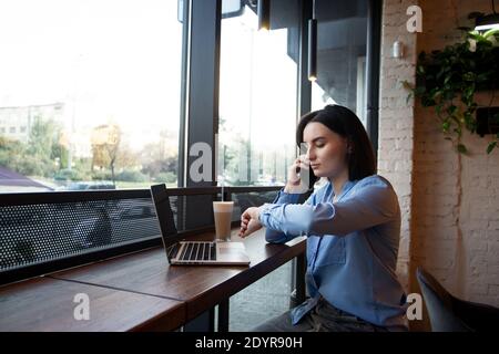 Busy freelancer woman looking at her watch and discussing something with customer. Remote work concept. Cafe or restaurant on background. Little time Stock Photo