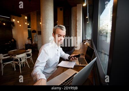 Freelance programmer sits near the window in a coffee shop and works. Stylish blonde man in white shirt uses laptops. Cozy coffee shop atmosphere Stock Photo