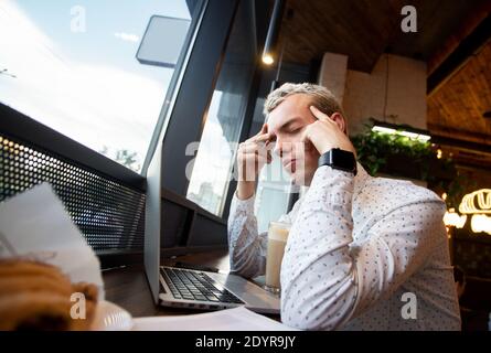 Freelance man having migraine at cafe. Man working in coffee house front of laptop suffering from chronic daily headaches. Young man holding painful Stock Photo