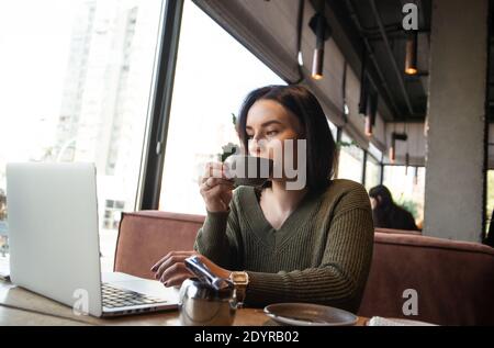 Freelancer working in cozy atmosphere concept. Concentrated young woman drinks coffee from grey cup and looks into laptop on her table with attention Stock Photo