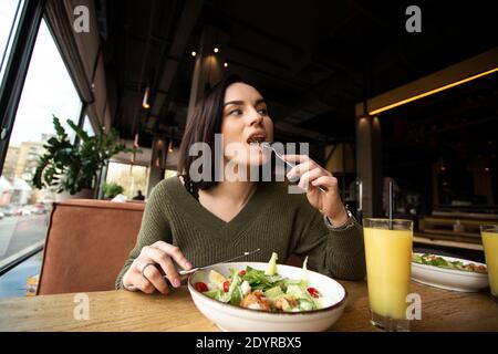 Young woman enjoys tasty meal. Attractive woman with brown hair slowly eating healthy caesar salad and looking away. Glass of orange juice. Restaurant Stock Photo