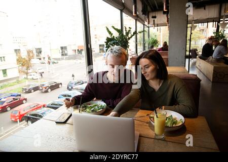 Couple spending time in restaurant together. Attractive young woman smiles and shows something on laptop to her boyfriend. They eat salads and drink Stock Photo