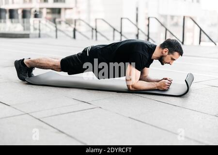 Plank it Training in big city concept. Sporty young man wearing black sportswear and doing plank position while exercising on asphalt roads. Hard Stock Photo