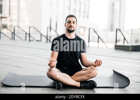 Man practicing yoga in big city. Sporty man meditating after workout. Excercising at home. Young man with stubble sitting on black yoga mat with eyes Stock Photo