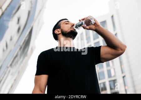 Athletic man drinking water from plastic bottle after jogging. Sporty male runner with dark short hair. Black t-shirt. Blurred apartment blocks on Stock Photo
