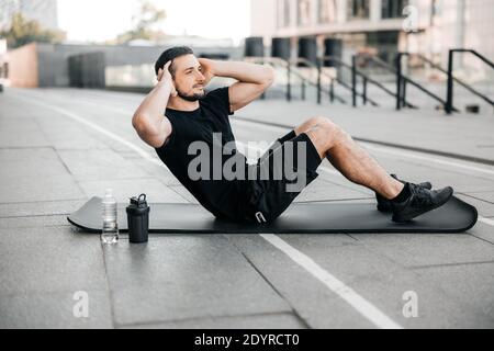 Fit man pumping press lying on black yoga mat. Muscular man doing his favorite morning exercise. Black sport clothes. Fit happy man doing fitness Stock Photo