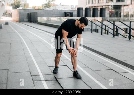 Attractive man feeling pain in his foot during sport workout outdoors. Male runner in black sportswear and sneakers standing and feeling cramps in his Stock Photo