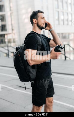 Friendly man in black clothes talking on the phone, smiling and looking away. Man standing near apartment blocks, holding black sports bottle. Urban Stock Photo
