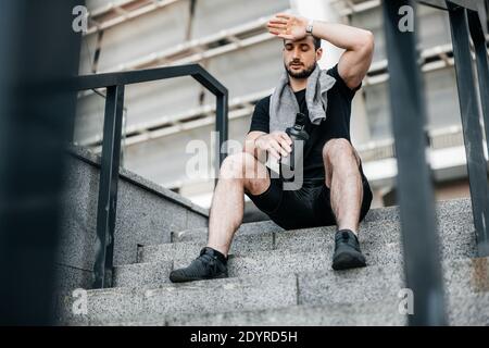 Resting time A man in black clothes with towel on his shoulders sits at the top of the stairs and rests after a hard jog or workout and wipes sweat Stock Photo