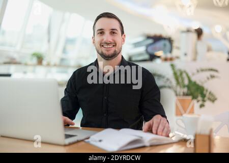 Cheerful freelancer man working from cafe. Attractive young man smiling, using his modern laprtop and looking at camera. Cozy coffeehouse on Stock Photo