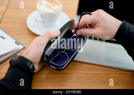 Take care of your eyeglasses. End of shift at remote work. Young man hides glasses in case. Male freelancer works from coffee shop, drinks flat white Stock Photo