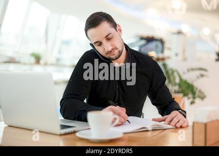 Businessman writes date of meeting with client in his planner. Young man in black shirt working from coffeehouse, drinking cappuccino and using his Stock Photo