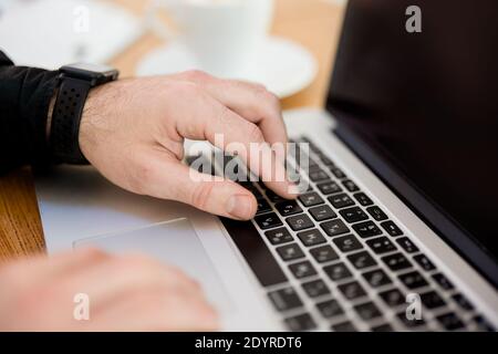 Male hands typing on laptop keyboard. Cut view. Modern gadgets. Close up. Gray laptop. Blurred cup of coffee on background. Man in black shirt. Male Stock Photo