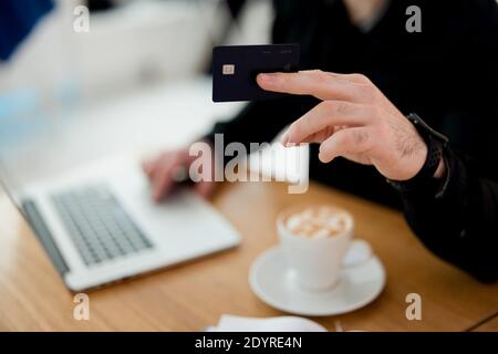 Focus on black credit card. Man is ready to pay for his order in a coffee shop. He holds card to barista. Blurred male body in black shirt, laptop and Stock Photo