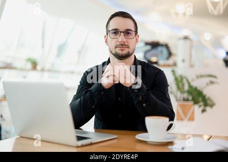 Confident attractive man looking at camera. Modern laptop and cup of cappuccino on wooden table. Light smart coffee shop interior on the background Stock Photo