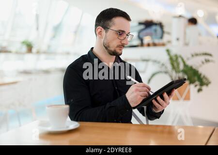 Male web designer or artist drawing on his tablet. Digital artwork concept. Young concentrated man with beard and eyeglasses using his gadget in Stock Photo