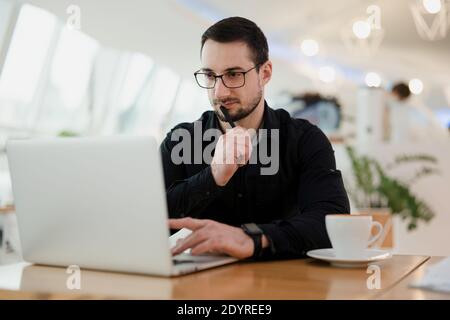 Concentrated man using laptop, holding pen near the mouth and thinking about his task. Work from cafe. Attractive intelligent man with beard wearing Stock Photo