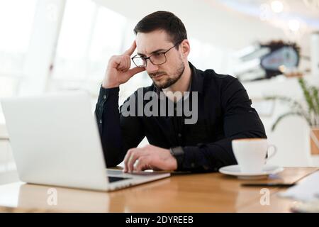 Smart young professional thinking about technical task. Concentrated man holding his head and using laptop. Remote work concept. Cup of yummy Stock Photo