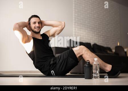 Sporty man pumping press lying on mat. Fit happy man doing fitness exercises on floor at home. Black sport clothes. Muscular man doing his favorite Stock Photo