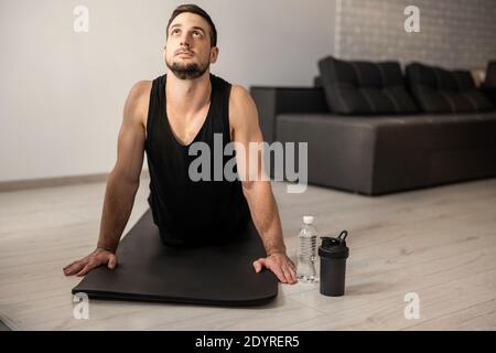 Fitn man meditating while doing cobra pose in living room. Indoor shot of handsome young man practicing yoga. Morning workout. Black sportswear Stock Photo