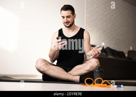 Man resting after home workout with water and phone. Young sporty man after practicing yoga, break in doing exercise, relaxing on yoga mat, texting on Stock Photo