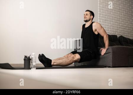 Attractive man pushing from sofa. Black sportswear. Man practicing hard to strengthen his arms. Plastic water bottle. Black yoga mat. Happy cheerful Stock Photo