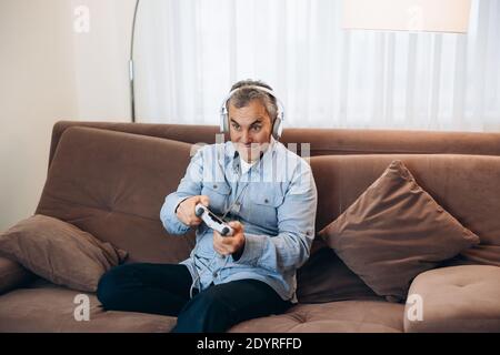 Adult man playing on console. Good time on weekend concept. Concentrated man with headphones sitting on brown sofa, holding game controller and Stock Photo