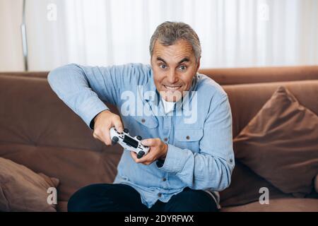 Impossible to put down Mature man is completely absorbed in playing his favorite game. He presses hard on buttons on game console. Living room on Stock Photo