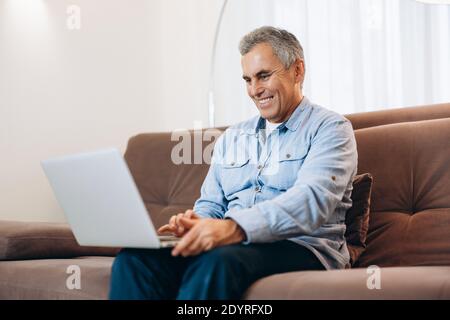 Cheerful man sitting on sofa, using laptop at home in cozy living room. Casual style. Middle aged man reads email, smiles and responds to it. Modern Stock Photo