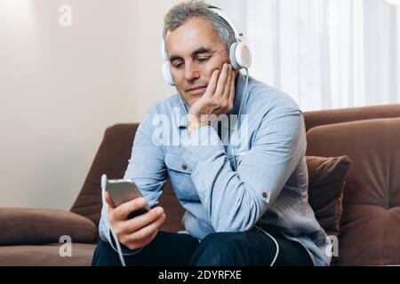 Senior man with headphones is watching an interesting movie from his phone. Entertainment during the lockdown. Mature man relaxes watching video on Stock Photo
