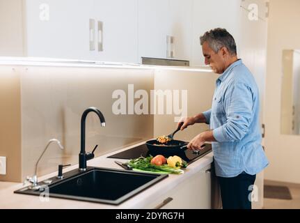Mature man preparing healthy food for dinner for his family. Concentrated man in casual clothes stands by the stove and stews vegetables in a frying Stock Photo