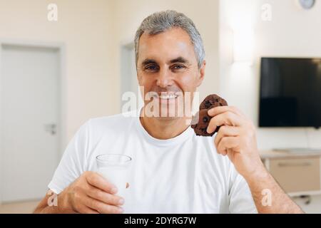 Adult man eating cookies and drinking milk. Modern light room on background. Good loking man looking at camera and smiling with teeth. Biscuits with Stock Photo